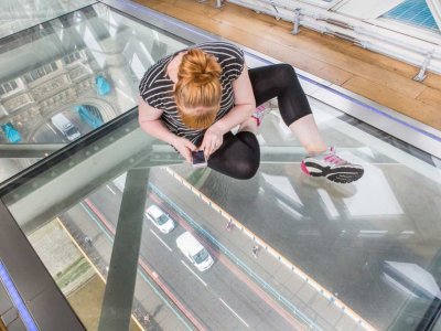 Woman sitting on Tower Bridge's Glass floor, looking through it at the road below.