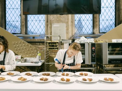 Two chefs in aprons are putting food onto a row of plates. 