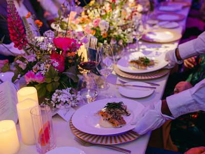 A long trestle table is laid with a white table cloth, flowers and candles. People are seated either side and servers are placing food in front of them. 