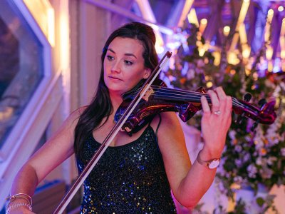 A violinist in a black sparkly dress plays while standing in the Tower Bridge Walkways with flowers and candles in the background.