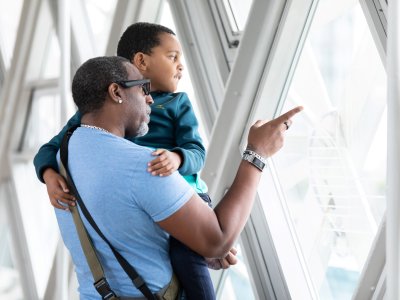 A man holding his child, both are looking out of the windows of the Tower Bridge Walkways at the view. The man is pointing to something out the window. 
