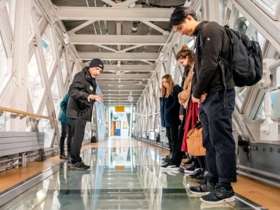 A group of visitors look down through the Glass Floor of the Tower Bridge Walkways, while a Visitor Host in black clothes gives a tour