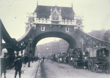 Historic photo of Tower Bridge in London around 1915, showing horse-drawn wagons and pedestrians crossing the bridge under its ornate stone arch.