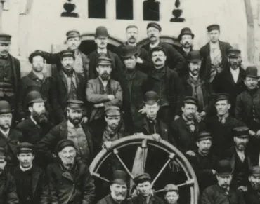 A black and white photo of a group of men wearing hard bowler hats and dark suits standing behind a large wooden wheel.