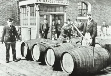 Victorian-era scene showing wooden cargo barrels arranged on a cobblestone street outside a vault entrance, with workers using long tools to handle the barrels.