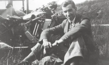 A black and white photo of a man in a suit sitting on grass in front of a motorbike, smiling at the camera.