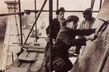 Historic photo of three workers on the roof of Tower Bridge engaged in construction atop scaffolding.