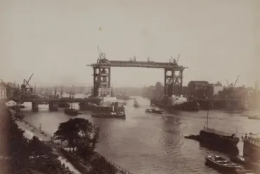 Historic photograph of Tower Bridge under construction in London, showing incomplete steel framework, cranes, and boats on the River Thames.
