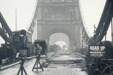 Historic black-and-white photo of Tower Bridge in London under road construction, featuring horse-drawn carts, early motor vehicles, and a sign reading 'Road Up' during infrastructure works.
