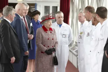 Royal visit inside Tower Bridge in London with Queen Elizabeth II, showing a group of officials and staff in suits and white uniforms gathered in a glass-panelled walkway with red curtains.