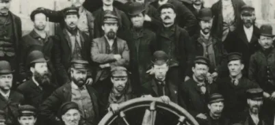 A black and white photo of a group of men wearing hard bowler hats and dark suits standing behind a large wooden wheel.