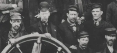 A black and white photo of a group of men wearing bowler hard-hats and suits. They are standing behind a a large wooden wheel.