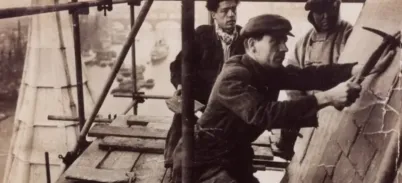 Historic photo of three workers on the roof of Tower Bridge engaged in construction atop scaffolding.