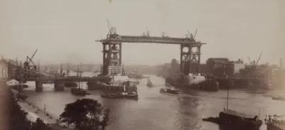 Historic photograph of Tower Bridge under construction in London, showing incomplete steel framework, cranes, and boats on the River Thames.