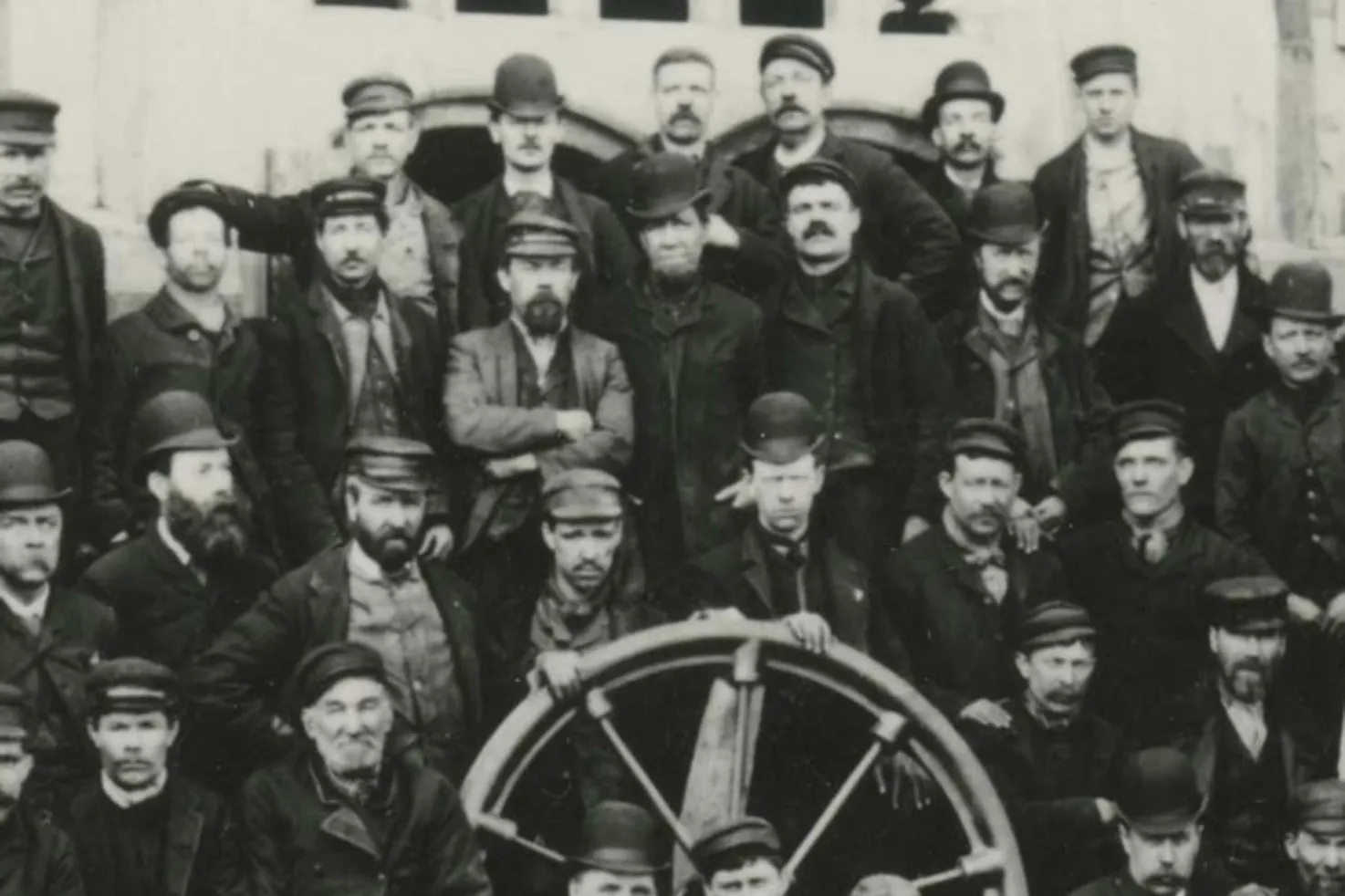 A black and white photo of a group of men wearing hard bowler hats and dark suits standing behind a large wooden wheel.