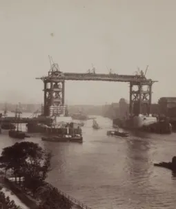 Historic photograph of Tower Bridge under construction in London, showing incomplete steel framework, cranes, and boats on the River Thames.