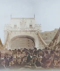 Historic photograph of a large crowd gathered on Tower Bridge during its public opening day in 1894, with the iconic Gothic-style towers and suspension chains prominently visible in the background.