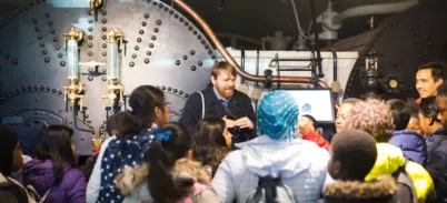 A group of schoolchildren look towards a tour guide standing in front of the Tower Bridge engine boilers