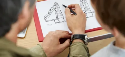 An adult hand is drawing a pen sketch of Tower Bridge on white paper.