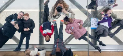 From above, eight children and adults lie on their backs on the glass floor of the Tower Bridge walkway, with the road and river just visible far below. In the middle the photographer is pointing a camera up at the ceiling mirror to take this image.