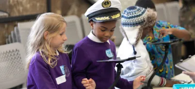 Two schoolchildren in purple jumpers look at an iPad. One wears a ship's captain's hat.