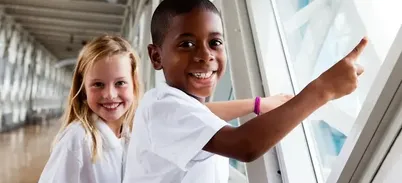 Two children in white school shirts are standing in the Walkway at the top of Tower Bridge, pointing out the window and smiling