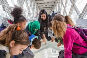 A group of children and one adult look and point down through the Tower Bridge Walkway Glass Floors