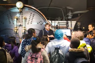 A group of schoolchildren look towards a tour guide standing in front of the Tower Bridge engine boilers