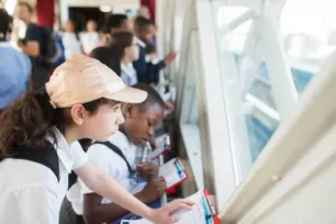 A child in a cap and other schoolchildren look at clipboards and out of the Tower Bridge walkway window.