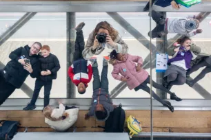 From above, eight children and adults lie on their backs on the glass floor of the Tower Bridge walkway, with the road and river just visible far below. In the middle the photographer is pointing a camera up at the ceiling mirror to take this image.