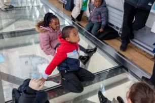 Two children sit on the Tower Bridge walkway glass floor, looking up at adults nearby