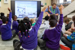 Schoolchildren in purple jumpers have their hands up, in front of a presentation screen and workshop leader.