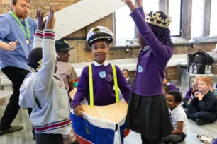 A child dressed as a boat with a captain's hat walks between two children with their hands raised to look like Tower Bridge. One wears a crown.