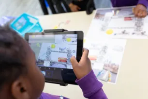 A schoolchild is holding a tablet, taking a photo of pictures of a bus and Tower Bridge on a table.