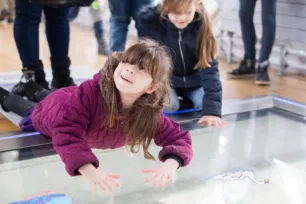 A girl in a purple jacket has her hands on the glass floor of a Tower Bridge walkway. She is looking up and smiling.