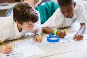 Two schoolchildren in white shirts write on worksheets, a blue magnifying glass lies between them.