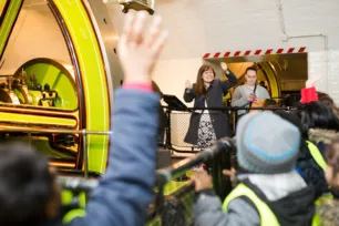 A group of schoolchildren with their hands up look towards a tour guide, who stands beside a green wheel of and engine.