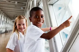Two children in white school shirts are standing in the Walkway at the top of Tower Bridge, pointing out the window and smiling