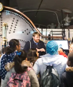 A group of schoolchildren look towards a tour guide standing in front of the Tower Bridge engine boilers