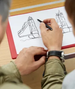 An adult hand is drawing a pen sketch of Tower Bridge on white paper.