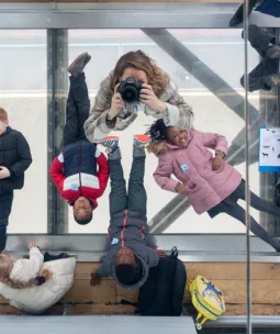 From above, eight children and adults lie on their backs on the glass floor of the Tower Bridge walkway, with the road and river just visible far below. In the middle the photographer is pointing a camera up at the ceiling mirror to take this image.