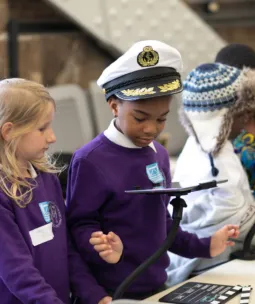 Two schoolchildren in purple jumpers look at an iPad. One wears a ship's captain's hat.
