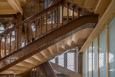 Interior metal staircase at Tower Bridge, London – featuring riveted steel steps, ornate wooden handrails, and industrial Victorian architecture illuminated by natural light through grid-patterned windows. The staircase is painted the original "bright chocolate" brown that once featured on the outside of Tower Bridge.