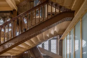 Interior metal staircase at Tower Bridge, London – featuring riveted steel steps, ornate wooden handrails, and industrial Victorian architecture illuminated by natural light through grid-patterned windows. The staircase is painted the original "bright chocolate" brown that once featured on the outside of Tower Bridge.