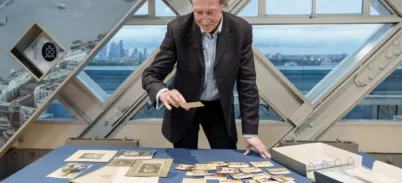 Person standing at a display table inside a glass-paneled structure, examining historical photographs and documents. The table is covered with vintage photos, papers, and an open binder. A large window behind reveals a cityscape with modern buildings in the distance.