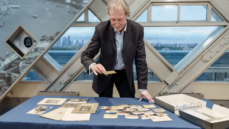 Person standing at a display table inside a glass-paneled structure, examining historical photographs and documents. The table is covered with vintage photos, papers, and an open binder. A large window behind reveals a cityscape with modern buildings in the distance.
