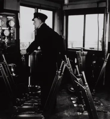 A Bridge Driver standing in the old Control Room over several large levers wearing a cap. 
The photo is black and white.