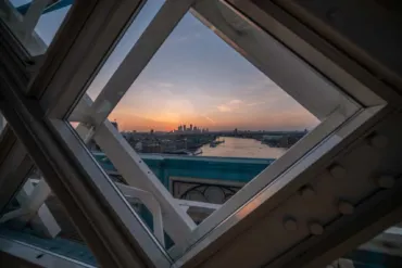 A diamond shaped window in the trelice like structure on the High-Level Walkways. Through it there is a city skyline and sunrise.