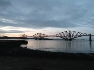 Forth Bridge at dusk – iconic red cantilever railway bridge in Scotland spanning the Firth of Forth, with steel triangular framework reflected in calm waters and a rocky shoreline in the foreground.