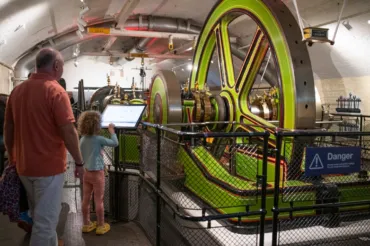 A man and young girls look at a large and brightly painted fly-wheel